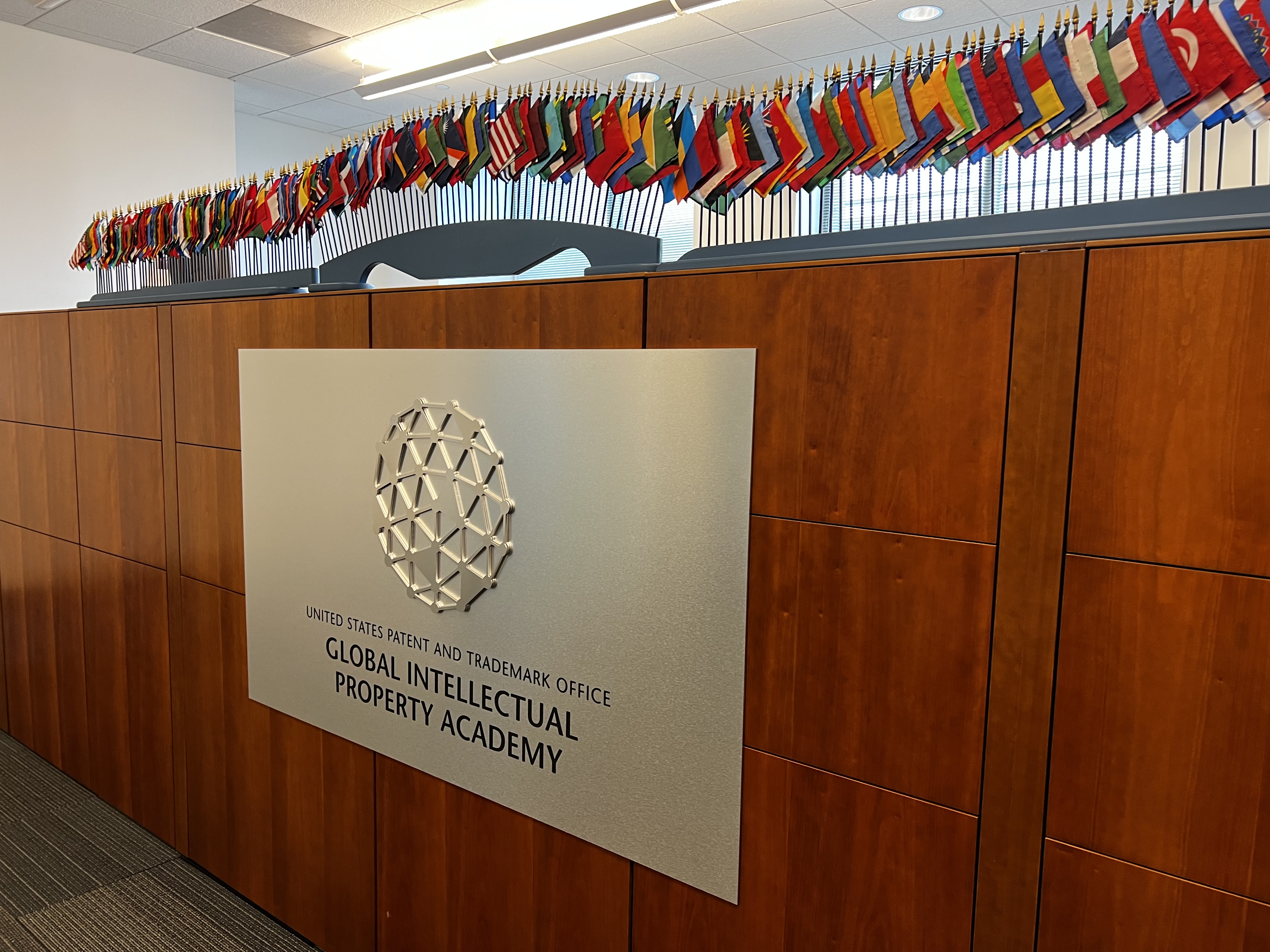 Silver sign on wooden wall reading "United States Patent and Trademark Office Global Intellectual Property Academy" underneath small flags. 