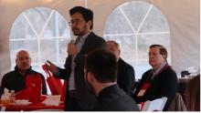 a graduate student holding a microphone and addressing a dinner crowd under a canopied tent.